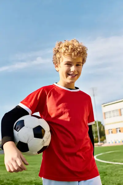 Молодой мальчик уверенно стоит на пышном зеленом футбольном поле, держа футбольный мяч с решимостью. Солнце светит ярко, бросая теплый свет на его страстное лицо. — стоковое фото
