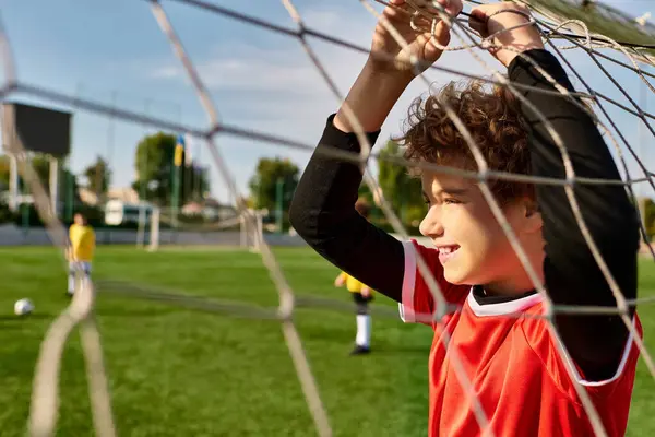 Un niño está detrás de una red de fútbol, sosteniendo una pelota de fútbol en sus manos. Su mirada enfocada sugiere determinación y pasión por el deporte mientras practica sus habilidades de portero. - foto de stock