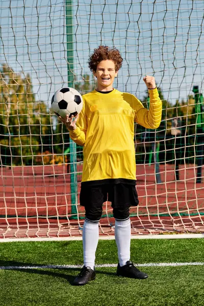Un jeune garçon en uniforme de football se tient debout avec confiance, tenant un ballon de football avec un regard déterminé sur son visage. — Photo de stock