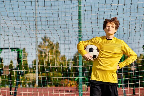 Un joven se para frente a una red de fútbol, sosteniendo una pelota de fútbol. Sus ojos están enfocados, listos para asumir el reto de marcar un gol. El campo verde se extiende detrás de él, bajo un cielo azul claro. - foto de stock