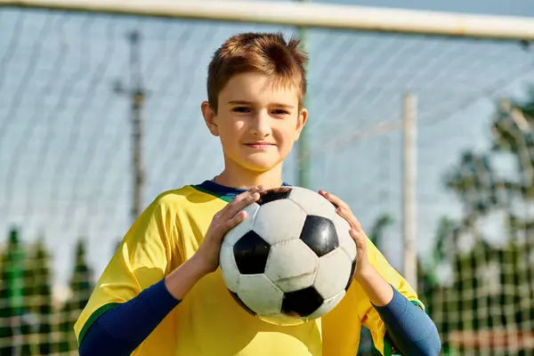 Um menino fica na frente de um gol de futebol, segurando uma bola de futebol em suas mãos. Ele olha para a frente com determinação, pronto para dar um tiro em direção à rede. — Fotografia de Stock