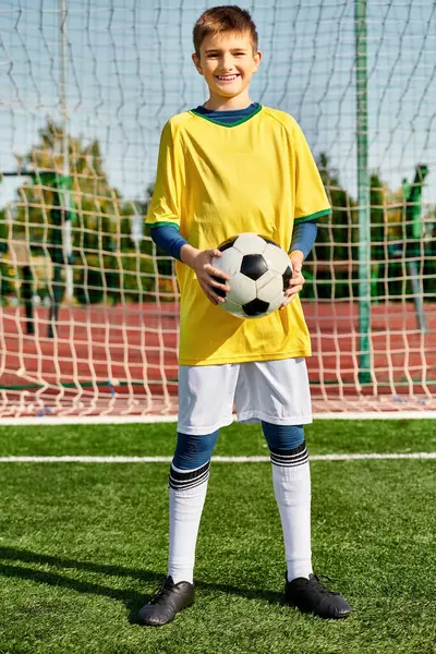 Une scène vibrante d'un jeune garçon debout sur un terrain de football vert luxuriant, tenant un ballon de football. Ses yeux brillent d'excitation alors qu'il se prépare à donner un coup de pied au ballon, exsudant une passion pour le sport. — Photo de stock