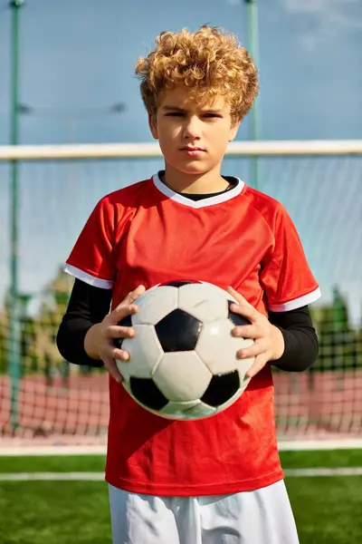 Un jeune garçon se tient en confiance sur un vaste terrain de soccer, berçant un ballon de soccer près de sa poitrine. L'herbe vert vif s'étend autour de lui, sous un ciel bleu clair. Ses yeux scintillent de détermination et d'excitation alors qu'il envisage le jeu à venir. — Photo de stock