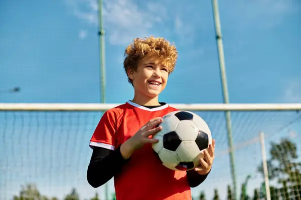 Um menino fica na frente de um gol de futebol, segurando uma bola de futebol com uma expressão determinada. Ele está posicionado para um chute, mostrando seu amor pelo esporte e sua prontidão para marcar um gol. — Fotografia de Stock