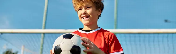 Un niño está parado en un vasto campo verde, agarrando una pelota de fútbol con determinación. Él mira hacia adelante, listo para patear la pelota y jugar un juego animado. - foto de stock