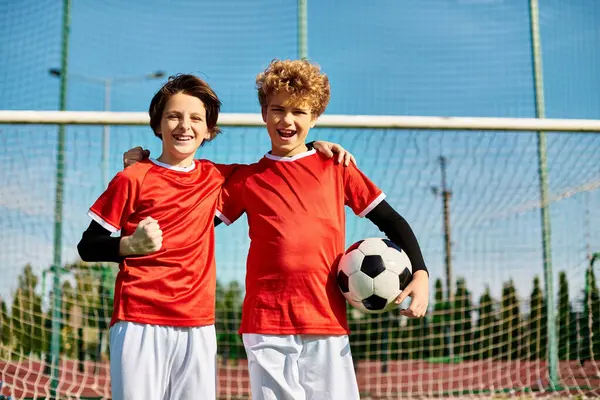 Двое детей стоят бок о бок, держа футбольный мяч. Их выражения выражают волнение и решимость в ожидании совместной игры. Яркие цвета их майки добавляют динамичной и энергичной атмосфере. — стоковое фото
