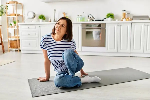 Una bella donna matura in accoglienti casalinghe che praticano yoga su un tappeto in una cucina illuminata dal sole. — Foto stock