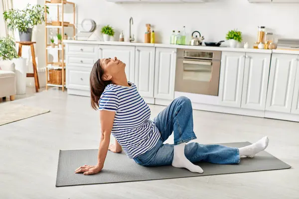 Una mujer madura en ropa acogedora practica yoga en una alfombra de cocina. - foto de stock
