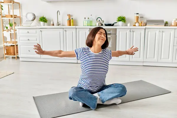 Mature woman in cozy homewear sitting on a yoga mat, meditating in a sunlit kitchen. — Stock Photo