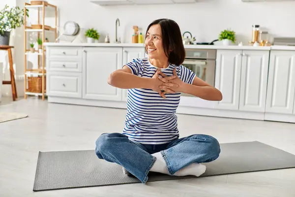 Зріла красива жінка в затишному домашньому вбранні практикує йогу на килимку на своїй кухні. — стокове фото