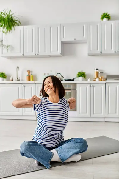 Una mujer madura en acogedora ropa de casa practica yoga sobre una alfombra en su cocina. - foto de stock