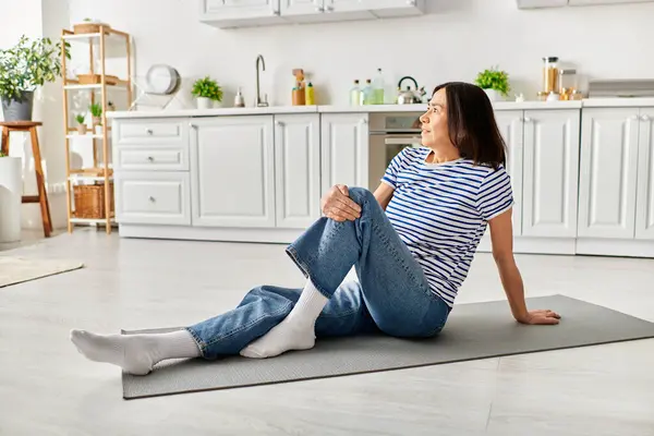 Mujer madura en acogedora ropa de casa encuentra la paz en la esterilla de yoga en la cocina. - foto de stock