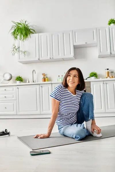 Una mujer madura y hermosa en ropa de casa acogedora practicando yoga en una alfombra en su cocina. - foto de stock
