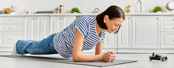 Una mujer madura en ropa de casa practica yoga en una alfombra de cocina. - foto de stock