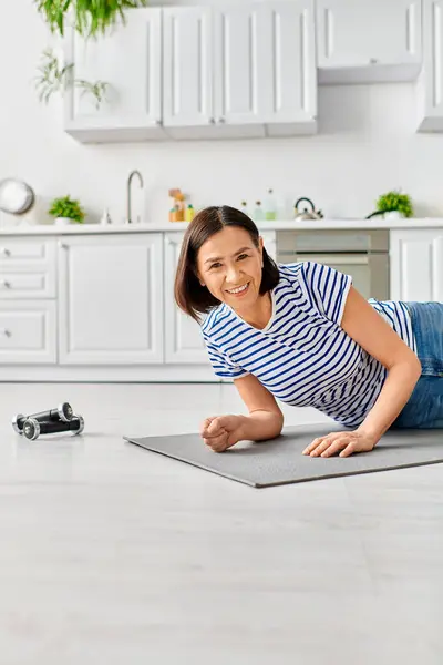 A mature woman in homewear practices yoga on a mat in a sunlit kitchen. — Stock Photo
