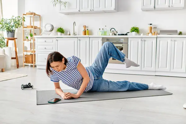 Mature woman in cozy attire practicing yoga on a mat in a home kitchen setting. — Stock Photo