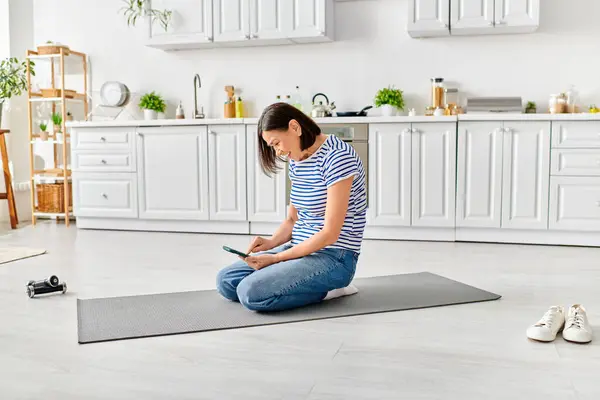 Mature woman in cozy attire sits on yoga mat, using cell phone in serene morning space. — Stock Photo