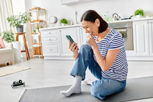 Uma mulher madura e bonita em roupa de casa aconchegante senta-se no chão, absorvida em seu telefone celular. — Fotografia de Stock