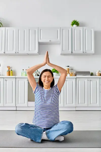 Mature woman in cozy homewear, sitting on a yoga mat with hands in the air. — Stock Photo