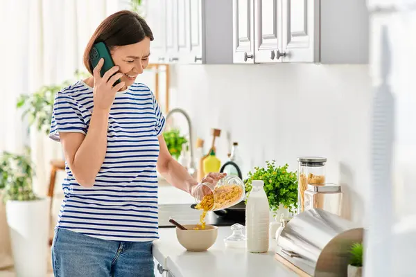 Une femme mûre en tenue de maison bavardant sur un téléphone portable alors qu'elle se tenait dans une cuisine. — Photo de stock