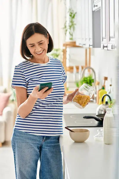 Mature woman in cozy homewear holding cell phone in kitchen. — Stock Photo