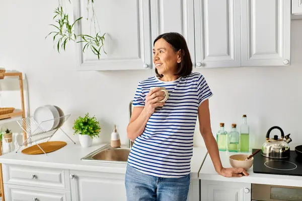 A mature woman in cozy homewear stands by the kitchen sink. — Stock Photo