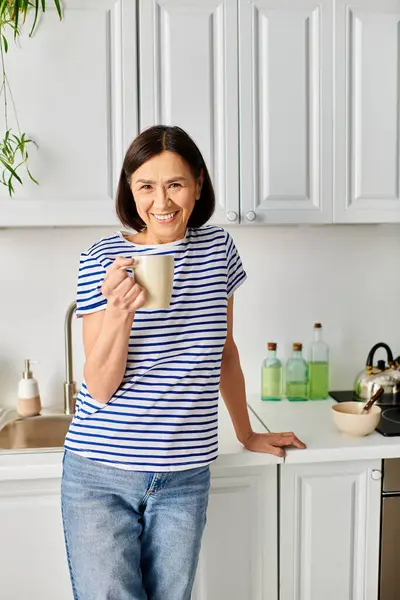 Una mujer en ropa de casa acogedora está en una cocina, sosteniendo una taza. - foto de stock