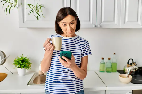 A woman in cozy homewear standing in a kitchen, holding a cup and a cell phone. — Stock Photo