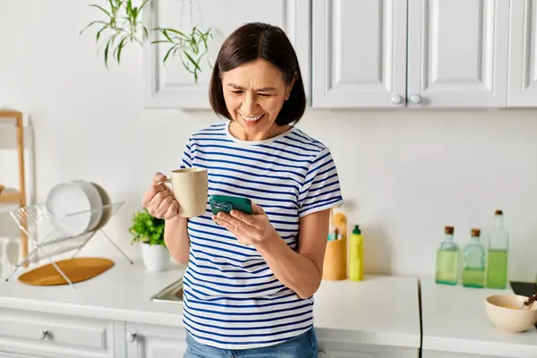 Una mujer en ropa de casa acogedora sosteniendo una taza, absorta en su teléfono celular. - foto de stock