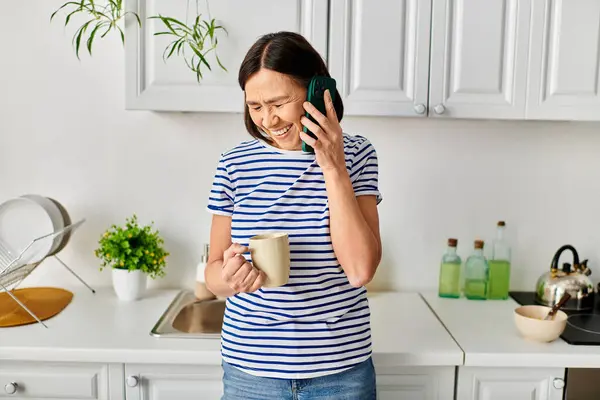 A mature woman in cozy homewear standing in a kitchen, chatting on a cell phone. — Stock Photo