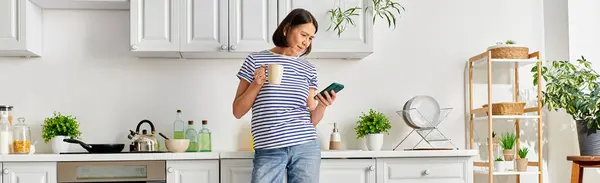 A mature woman in cozy homewear stands in a kitchen, holding a cell phone. — Stock Photo