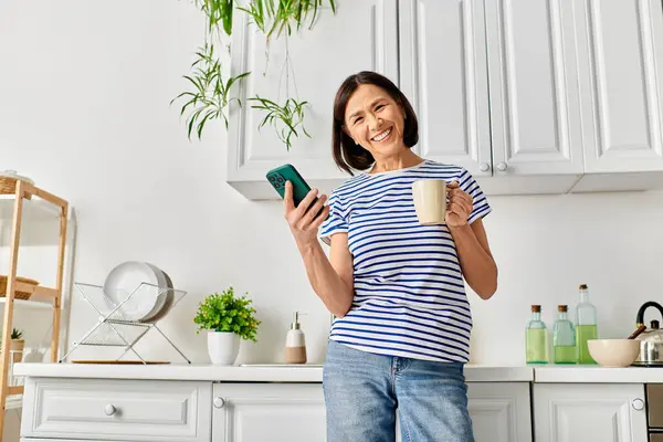 Mujer en acogedora ropa de casa taza de celebración y teléfono celular en la cocina. - foto de stock
