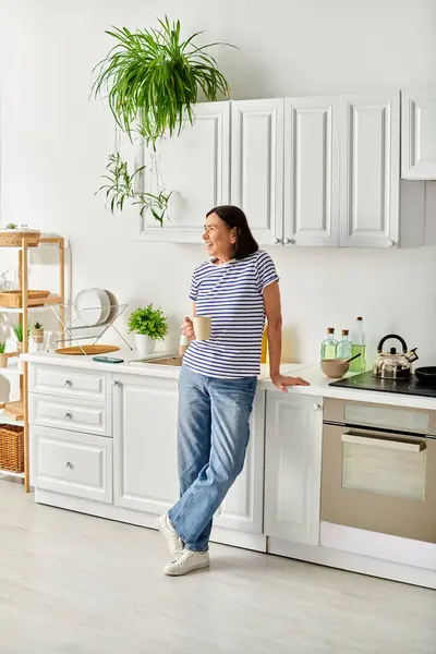 A mature woman in cozy homewear standing in a kitchen, next to a sink. — Stock Photo