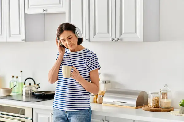 Una mujer madura en ropa de casa acogedora hablando en un teléfono celular en una cocina. - foto de stock