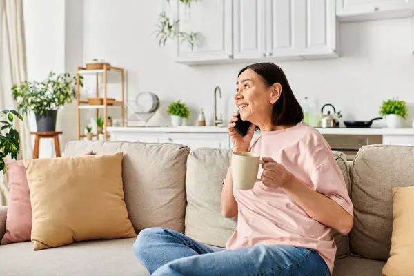 Una mujer madura en ropa de casa acogedora disfrutando de una taza de café mientras está sentada en un sofá. - foto de stock