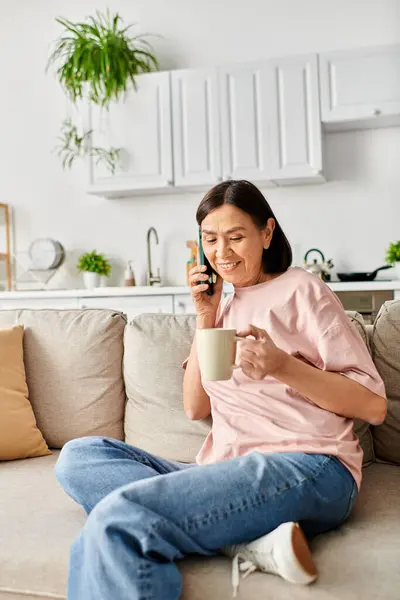 Una mujer madura en acogedora ropa de casa chats en un teléfono celular mientras se relaja en un sofá. - foto de stock