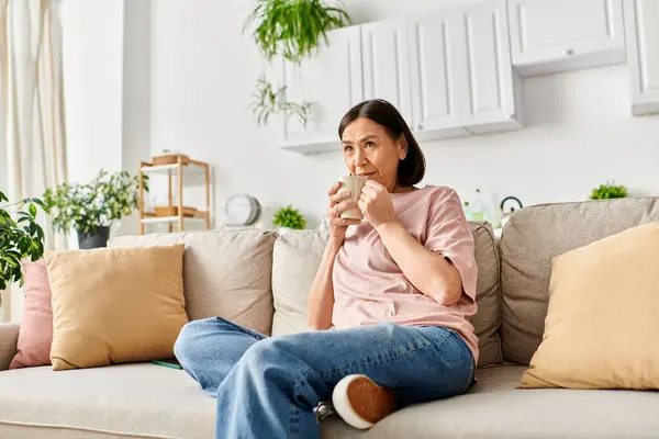 A mature woman in casual attire sits on a couch, happily eating a snack. — Stock Photo