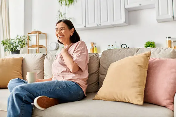 A mature woman in cozy homewear sitting on a couch, enjoying a cup of coffee. — Stock Photo