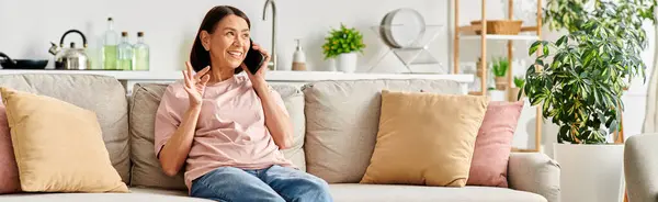 Una mujer madura en ropa de casa se sienta en un sofá, se dedica a una conversación telefónica. - foto de stock