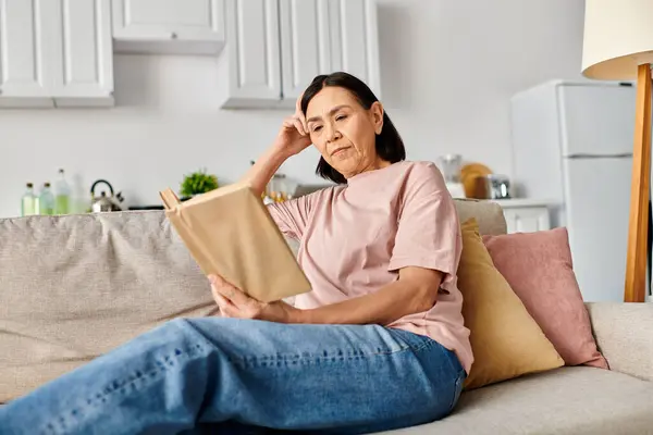A mature woman in cozy homewear sits on a couch, engrossed in reading a book. — Stock Photo