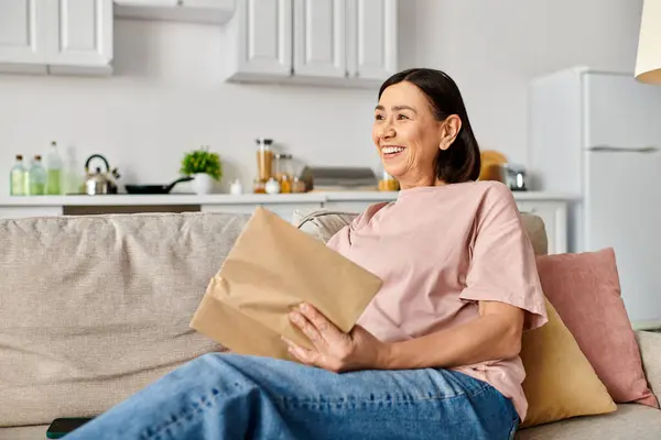 Una mujer madura en ropa de casa se sienta en un sofá, sosteniendo una bolsa de papel marrón. - foto de stock