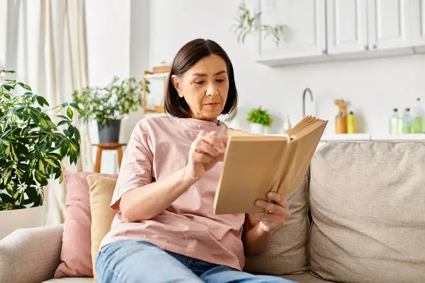 Mature woman in homewear seated on a couch, engrossed in reading a book. — Stock Photo