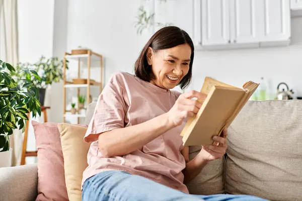 A mature, appealing woman in homewear, lost in the pages of a book while seated on a comfortable couch. — Stock Photo