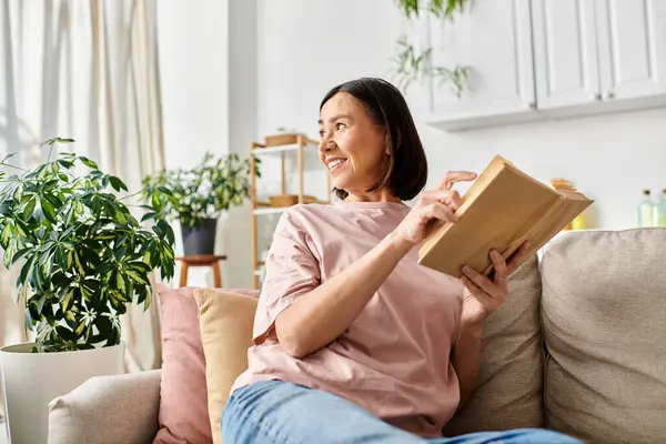 Зріла жінка в домашньому вбранні читає книгу зручно на дивані вдома. — стокове фото