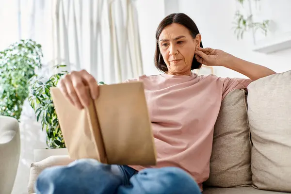 Eine reife Frau in bequemer Homewear sitzt auf einer Couch und ist in ein Buch vertieft. — Stockfoto