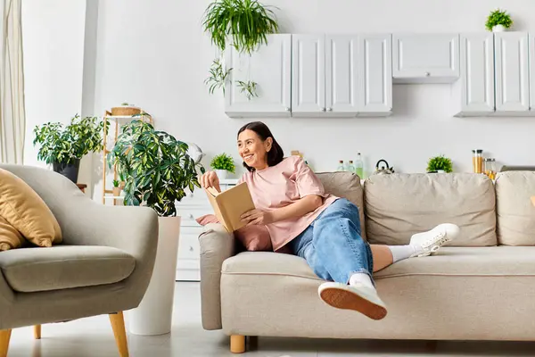 Eine Frau in gemütlicher Hausmannskleidung sitzt auf einer Couch, in ein Buch vertieft. — Stockfoto