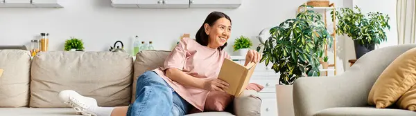 A mature woman in comfortable homewear sitting on a couch, lost in a book. — Stock Photo