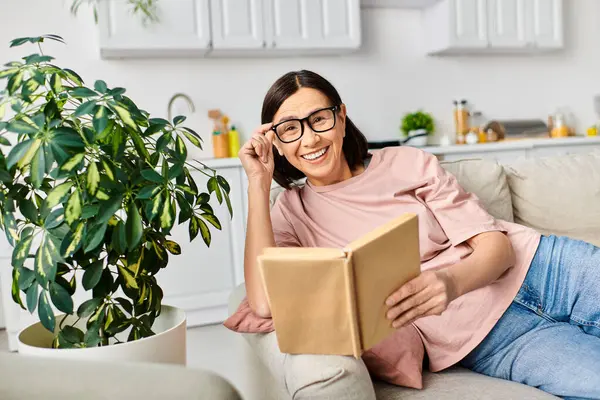 A mature woman in cozy homewear relaxes on a couch, immersed in a book. — Stock Photo