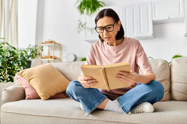 Eine reife Frau in kuscheliger Homewear sitzt auf einer Couch und liest ein Buch. — Stockfoto