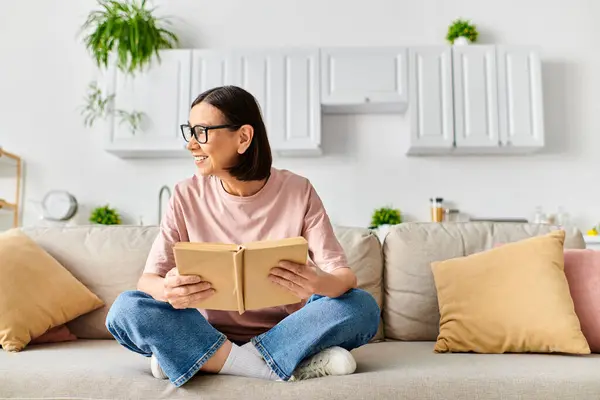 Женщина в уютной домашней одежде, погруженная в книгу, сидя на плюшевом диване. — стоковое фото